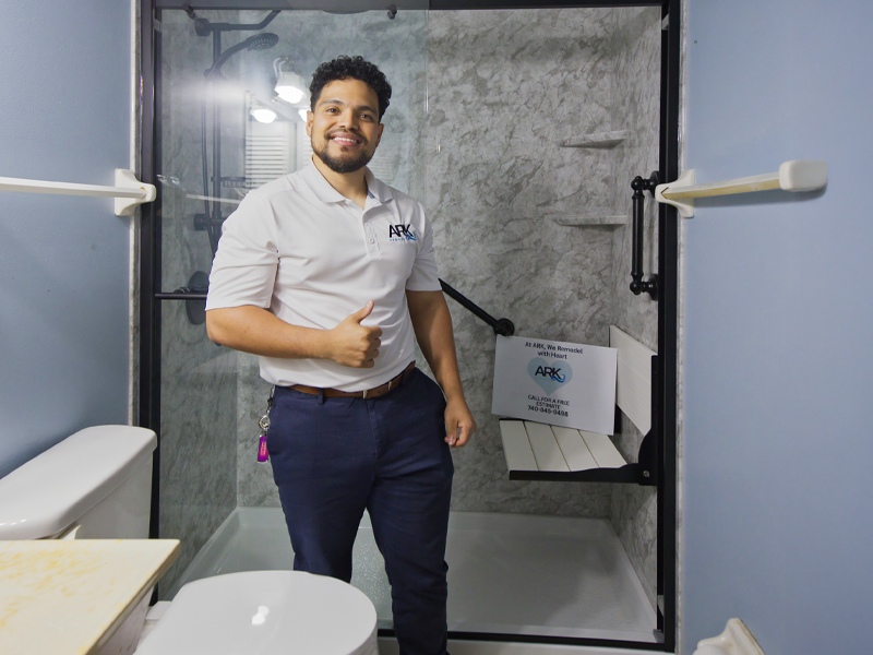ARK Remodel owner standing in a finished walk-in shower with a fold-down seat and grab bars