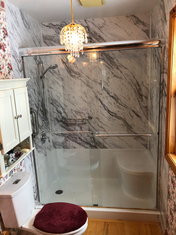 Walk-in shower with a moulded corner bench seat, grab bar and marble-look wall panels behind sliding glass doors